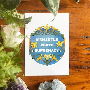 May include: A white card with a blue circle and yellow flowers. The text "Dismantle White Supremacy" is written in black letters on the card.