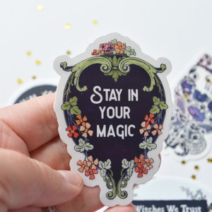 May include: A black and white sticker with a floral border and the text "Stay in your magic" in white lettering.