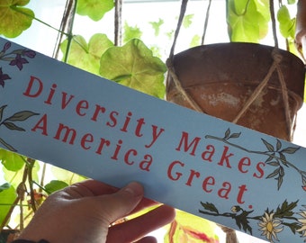 May include: A blue sign with the text "Diversity Makes America Great." in red lettering. The sign is decorated with floral illustrations in shades of red, orange, yellow, green, and purple.
