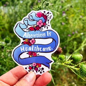Puede incluir: Una cinta azul con el texto "Abortion is Healthcare" en letras blancas. La cinta está decorada con flores rosas, rojas y amarillas. La pegatina se sostiene en una mano contra un fondo verde.