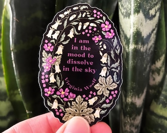 May include: A black and white oval sticker with pink flowers and white bells. The sticker has the text "I am in the mood to dissolve in the sky" and "Virginia Woolf" in a cursive font.