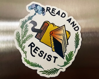 May include: A white circle sticker with green foliage around the edges. The sticker has the text "Read and Resist" in black lettering. A yellow and blue book with a red bookmark is in the center of the sticker.