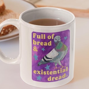 Puede incluir: Taza de cerámica blanca con el texto "Full of bread & existential dread" y un gráfico de paloma. La taza está sobre una bandeja blanca con dos rebanadas de pan y una manzana. Un ordenador portátil está en el fondo.