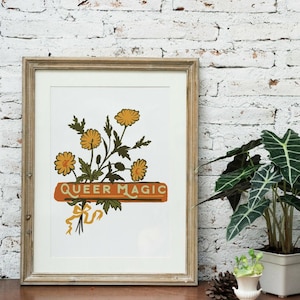 May include: A framed print with a white background featuring a bouquet of yellow flowers and the text "QUEER MAGIC" in orange and white lettering.