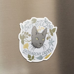 Può includere: Un magnete da frigorifero rotondo bianco con un'illustrazione di un gatto grigio e il testo "Cats Rule and Patriarchy Drools".
