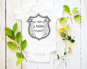 May include: White baby onesie with a black and white graphic of a shield with the text "My Mom is a Raging Feminist".