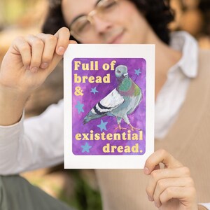 K&ouml;nnte beinhalten: Ein rechteckiger Kunstdruck mit einem Taubenmotiv vor einem violetten Aquarellhintergrund mit hellblauen Sternen. Der Text lautet Full of bread & existential dread in gelber Schrift. Die Taube hat graue, wei&szlig;e und schillernd gr&uuml;ne Federn.