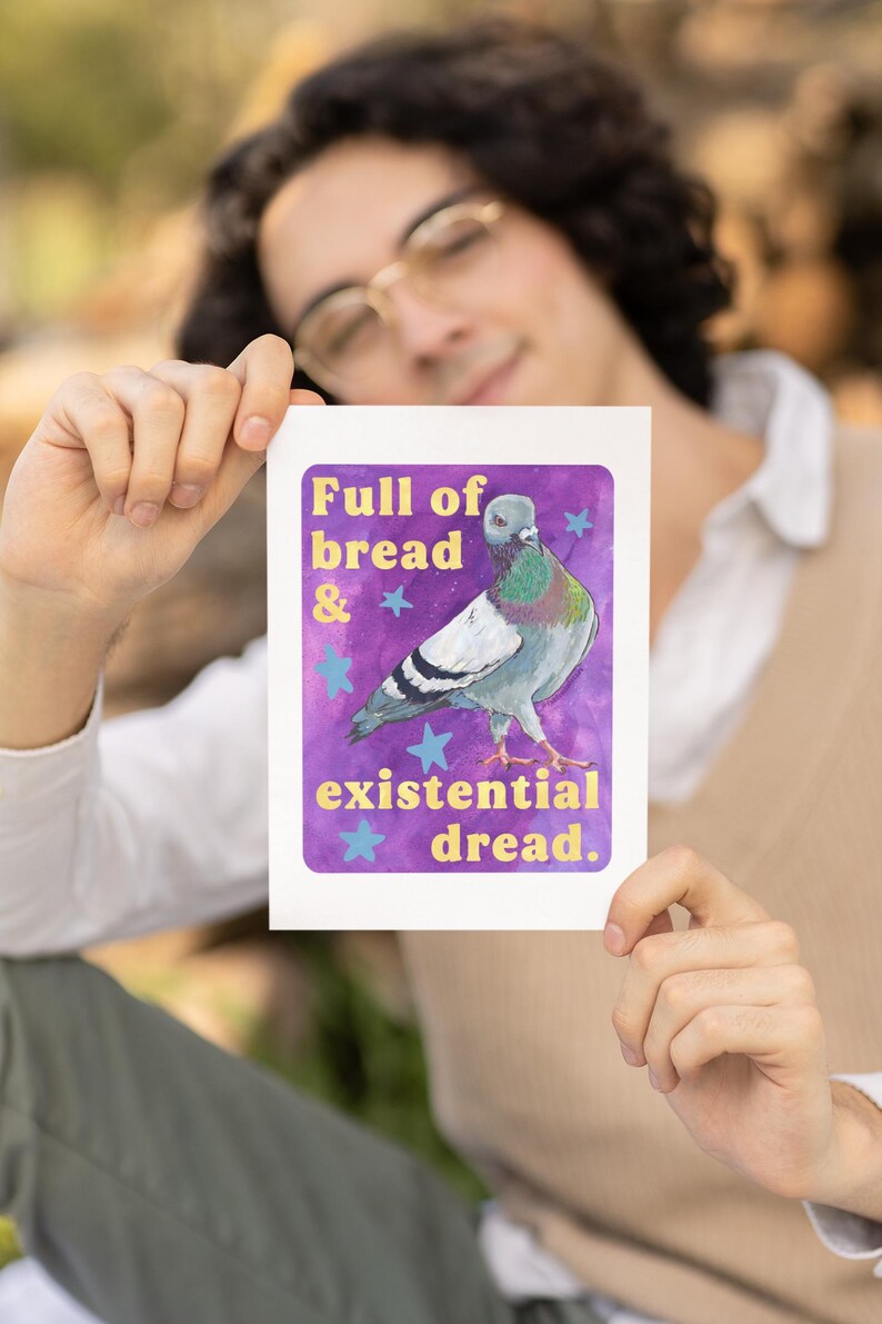 K&ouml;nnte beinhalten: Ein rechteckiger Kunstdruck mit einem Taubenmotiv vor einem violetten Aquarellhintergrund mit hellblauen Sternen. Der Text lautet Full of bread & existential dread in gelber Schrift. Die Taube hat graue, wei&szlig;e und schillernd gr&uuml;ne Federn.
