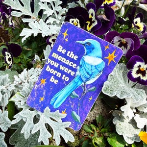 May include: A rectangular sticker with a blue bird illustration and the text "Be the menace you were born to be." The sticker is surrounded by purple pansies, silver foliage, and other greenery.