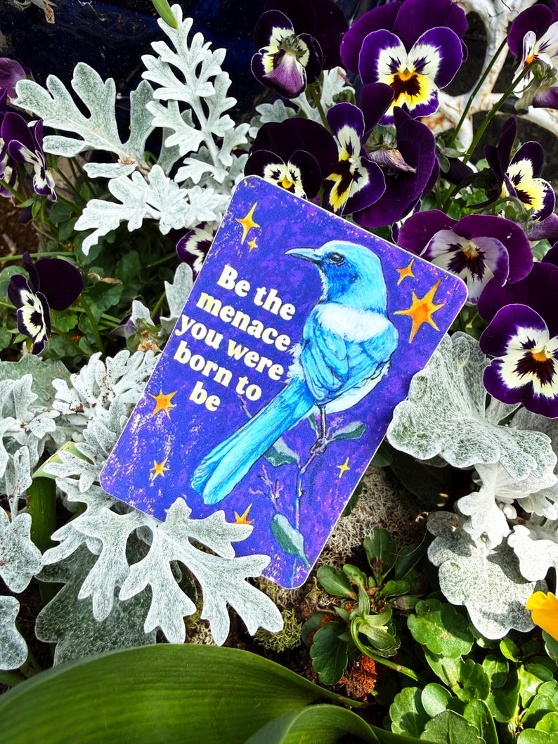 May include: A rectangular sticker with a blue bird illustration and the text "Be the menace you were born to be." The sticker is surrounded by purple pansies, silver foliage, and other greenery.