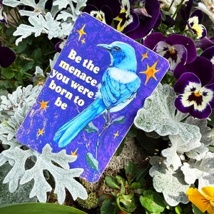 May include: A rectangular card with a blue bird illustration and the text "Be the menace you were born to be" is surrounded by purple pansies, silver foliage, and green plants.