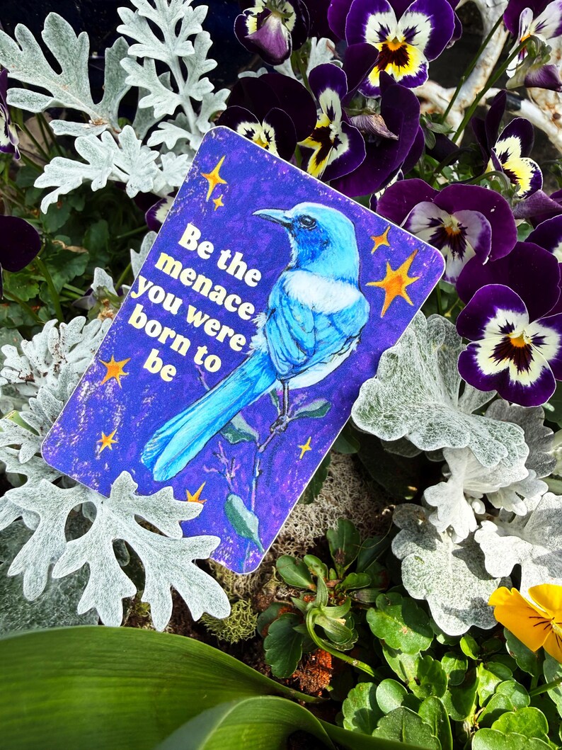 May include: A rectangular card with a blue bird illustration and the text "Be the menace you were born to be" is surrounded by purple pansies, silver foliage, and green plants.