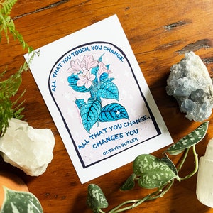 Peut inclure: Une carte blanche avec une illustration de fleur rose et du texte bleu qui dit "Tout ce que vous touchez, vous changez. Tout ce que vous changez, vous change. Octavia Butler."