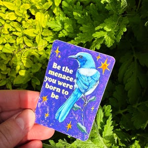 May include: A rectangular sticker with a blue bird illustration on a purple background, surrounded by yellow stars. The sticker has the text "Be the menace you were born to be". The sticker is held against a backdrop of green foliage.