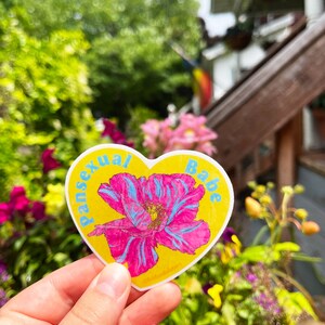 May include: A yellow heart-shaped sticker with the words "pansexual babe" in blue lettering. A pink flower with blue veins is in the center of the sticker.