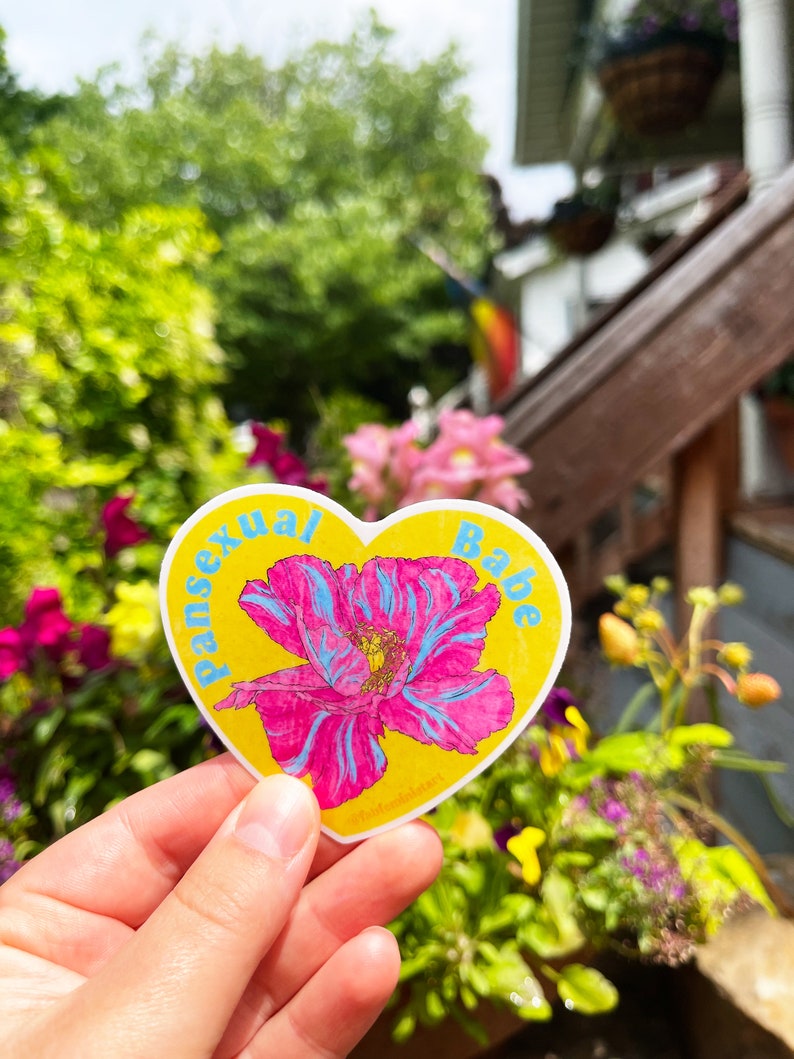 May include: A yellow heart-shaped sticker with the words "pansexual babe" in blue lettering. A pink flower with blue veins is in the center of the sticker.