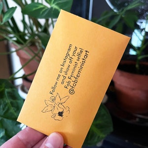 Puede incluir: Un sobre amarillo con una ilustraci&oacute;n en blanco y negro de una flor. El sobre tiene texto que dice "&iexcl;S&iacute;gueme en Instagram y presume de tu selfie feminista fabuloso! @fcbfeministart"