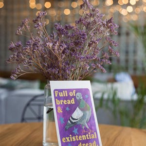 K&ouml;nnte beinhalten: Eine klare Glasvase gef&uuml;llt mit violetten getrockneten Statice-Blumen, platziert hinter einer rechteckigen Karte mit einer Taubenillustration auf violettem Grund mit blauen Sternen und dem Text Full of bread & existential dread.