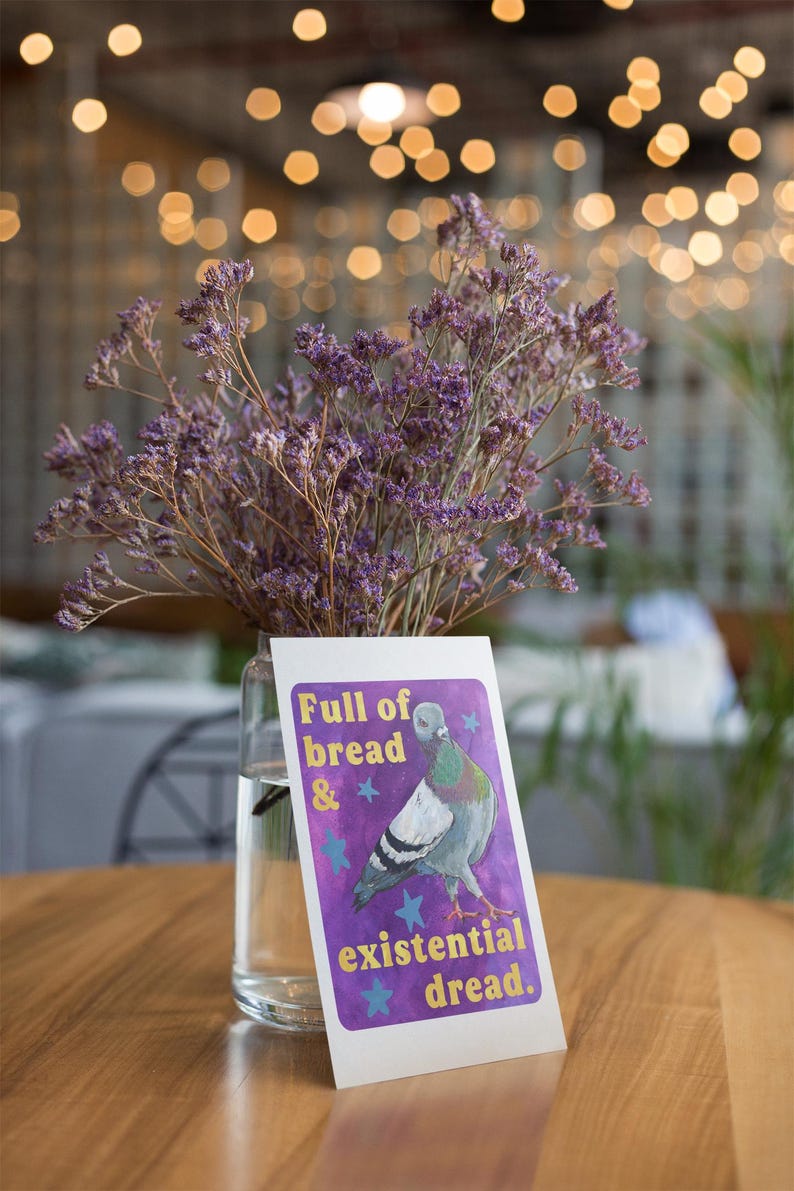 K&ouml;nnte beinhalten: Eine klare Glasvase gef&uuml;llt mit violetten getrockneten Statice-Blumen, platziert hinter einer rechteckigen Karte mit einer Taubenillustration auf violettem Grund mit blauen Sternen und dem Text Full of bread & existential dread.
