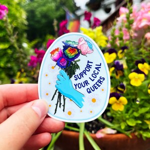 May include: A blue and white oval sticker with a rainbow rose bouquet, a blue gloved hand holding the stems, and the text "Support Your Local Queers".