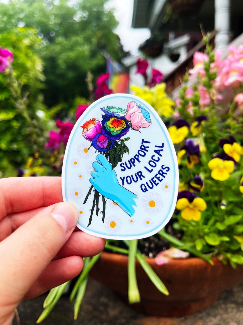 May include: A blue and white oval sticker with a rainbow rose bouquet, a blue gloved hand holding the stems, and the text "Support Your Local Queers".