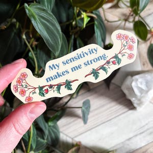 May include: A white sticker with a floral design and the text "My sensitivity makes me strong." The sticker is held in a hand.