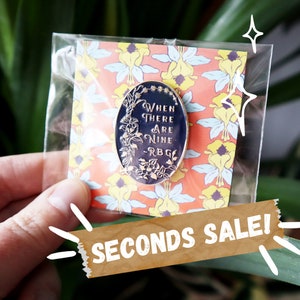Può includere: Uno spillo in smalto dorato con sfondo nero e testo bianco che recita "When there are nine - RBG". Lo spillo è circondato da un motivo floreale ed è in una busta di plastica trasparente.