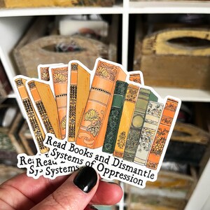 May include: A sticker with a stack of vintage-style books with ornate covers. The sticker has the text "Read Books and Dismantle Systems of Oppression" in black lettering.
