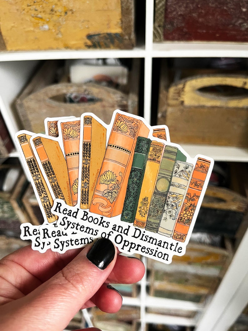 May include: A sticker with a stack of vintage-style books with ornate covers. The sticker has the text "Read Books and Dismantle Systems of Oppression" in black lettering.