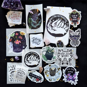 May include: A collection of stickers featuring feminist and witchy themes. The stickers are printed on white paper with black ink and feature various designs, including a hand holding a flame, a cat with the text "Patriarchy Drools", and a witch with the text "In Witches We Trust".
