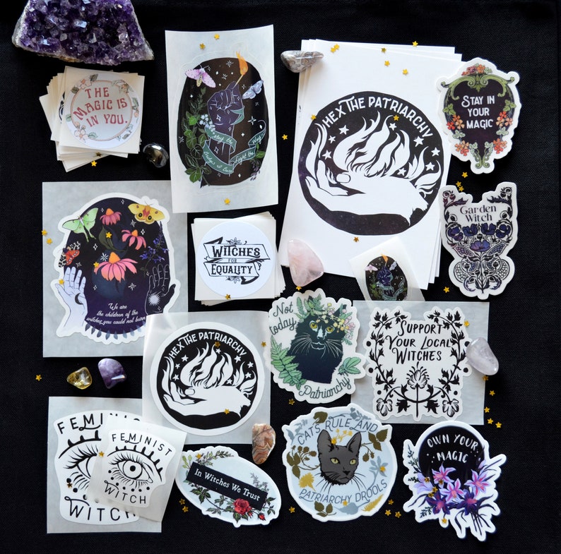 May include: A collection of stickers featuring feminist and witchy themes. The stickers are printed on white paper with black ink and feature various designs, including a hand holding a flame, a cat with the text "Patriarchy Drools", and a witch with the text "In Witches We Trust".