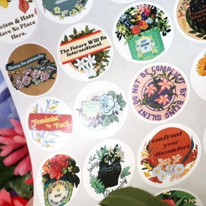 May include: A set of round stickers with various feminist slogans and floral designs. The stickers feature phrases like "Racism & Hate Have No Place Here", "The Future Will Be Intersectional", "Feminist AF", "Change Takes Courage", "Do Not Be Complicit By Being Silent", "The Fight Is Not Over", "Confront Your Discomfort", and "Another World Is Possible".