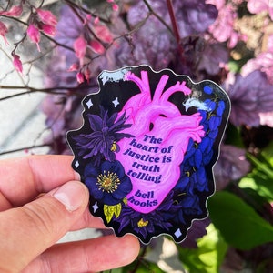 Puede incluir: Un coraz&oacute;n rosa con contorno negro y detalles florales. El coraz&oacute;n tiene el texto "The heart of justice is truth telling bell hooks" en su interior. La pegatina est&aacute; en una mano.