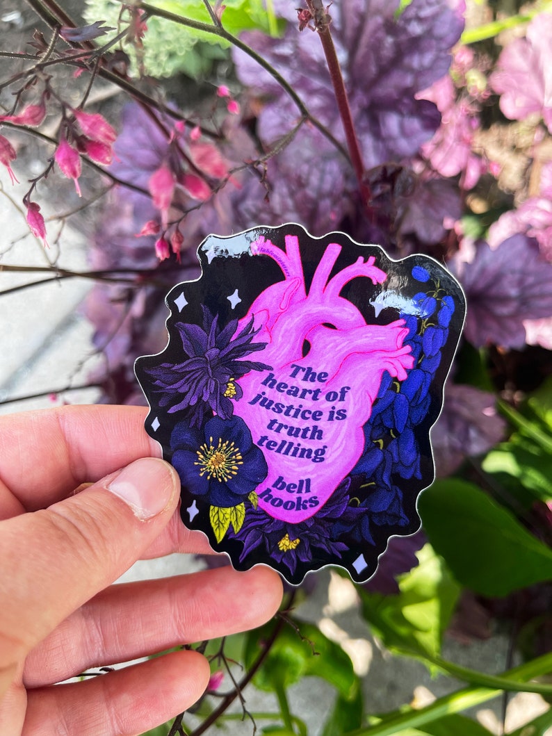 Puede incluir: Un coraz&oacute;n rosa con contorno negro y detalles florales. El coraz&oacute;n tiene el texto "The heart of justice is truth telling bell hooks" en su interior. La pegatina est&aacute; en una mano.