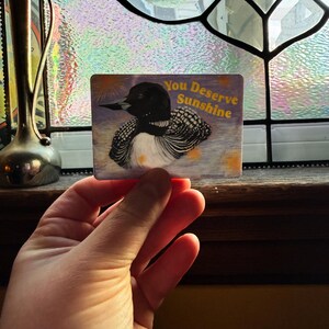 K&ouml;nnte beinhalten: Eine rechteckige Karte mit einer Abbildung eines Eistauchers, mit schwarzem Kopf und wei&szlig;er Brust. Die Karte tr&auml;gt den Text "You Deserve Sunshine" in Gelb. Der Hintergrund ist ein Bild im Aquarellstil.