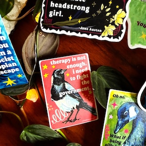 K&ouml;nnte beinhalten: Eine Sammlung bunter Aufkleber mit verschiedenen Designs und Zitaten. Ein Aufkleber zeigt eine Elster-Illustration mit dem Text "therapy is not enough. I need to fight the government." Ein anderer sagt "Obstinate headstrong girl."