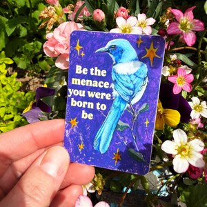 May include: A rectangular sticker with a blue bird illustration and the text "Be the menace you were born to be" on a dark blue background with yellow stars. The sticker is held in front of a background of colorful flowers.