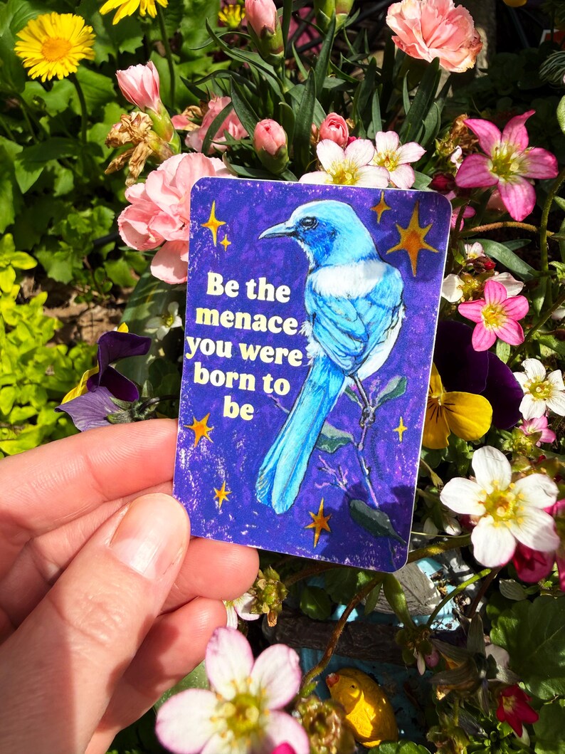 May include: A rectangular sticker with a blue bird illustration and the text "Be the menace you were born to be" on a dark blue background with yellow stars. The sticker is held in front of a background of colorful flowers.