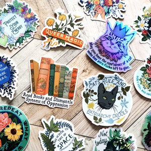 May include: A collection of colorful stickers with various feminist and LGBTQ+ themes. The stickers feature floral designs, cats, books, and text such as "Protect Your Energy", "Queer Magic", "Read Books and Dismantle Systems of Oppression", "All Bodies Are Good Bodies", and "Cats Rule and Patriarchy Drools".
