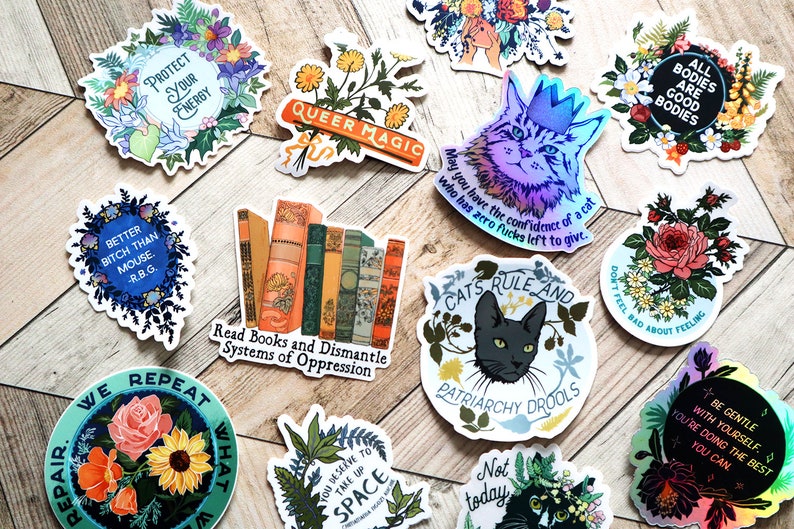 May include: A collection of colorful stickers with various feminist and LGBTQ+ themes. The stickers feature floral designs, cats, books, and text such as "Protect Your Energy", "Queer Magic", "Read Books and Dismantle Systems of Oppression", "All Bodies Are Good Bodies", and "Cats Rule and Patriarchy Drools".
