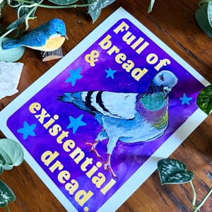 K&ouml;nnte beinhalten: Ein rechteckiger Kunstdruck mit einer Taube vor einem violett-blauen Galaxie-Hintergrund mit hellblauen Sternen. Der Text lautet Full of bread & existential dread in gelben Buchstaben. Die Taube hat graue, wei&szlig;e und schillernd gr&uuml;ne Federn.