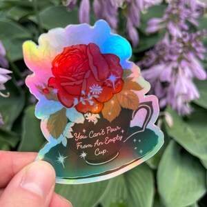 May include: A holographic sticker with a red rose in a teacup. The sticker has the text "You Can't Pour From An Empty Cup."