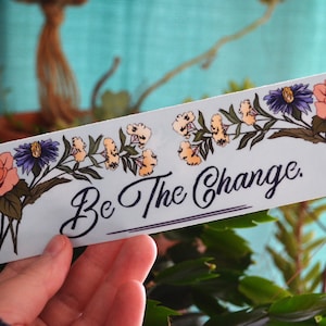 Puede incluir: Una pegatina rectangular con fondo azul claro, con ilustraciones florales y el texto "Be The Change". El diseño incluye rosas naranjas, flores moradas y amarillas, y hojas verdes.