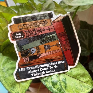 以下が含まれることがあります： 「Life-Transforming Ideas Have Always Come To Me Through Books」というテキストが書かれた、ビンテージな本の積み重ねが描かれたステッカーです。