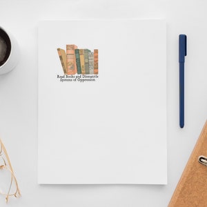 May include: A white notepad with a stack of vintage books and the text "Read Books and Dismantle Systems of Oppression" on a white surface. There is a cup of coffee, a pair of glasses, a blue pen, and a brown binder with metal rings.