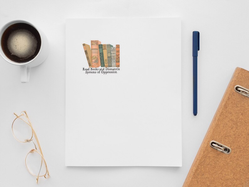 May include: A white notepad with a stack of vintage books and the text "Read Books and Dismantle Systems of Oppression" on a white surface. There is a cup of coffee, a pair of glasses, a blue pen, and a brown binder with metal rings.