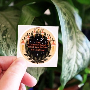 May include: A round sticker with a yellow background and a black circle in the centre. The black circle features a crescent moon, stars, and a floral design with red, orange, and yellow flowers. The text "You Cannot Change What You Refuse To Confront" is written in black on a white background.