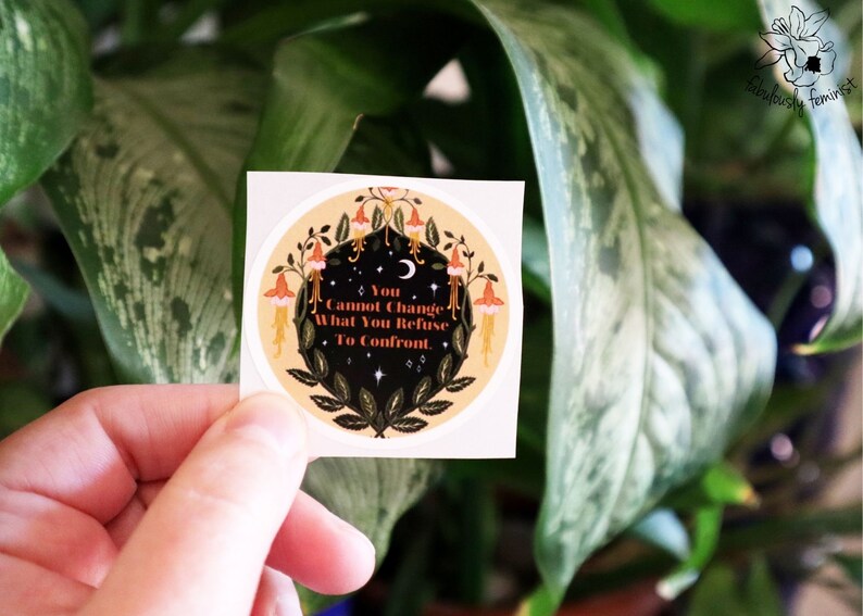 May include: A round sticker with a yellow background and a black circle in the centre. The black circle features a crescent moon, stars, and a floral design with red, orange, and yellow flowers. The text "You Cannot Change What You Refuse To Confront" is written in black on a white background.