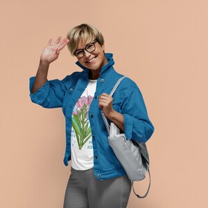 Puede incluir: Una mujer con una chaqueta vaquera azul sobre una camiseta blanca con estampado floral. Sonr&iacute;e y saluda con la mano izquierda. Lleva un bolso gris sobre el hombro.
