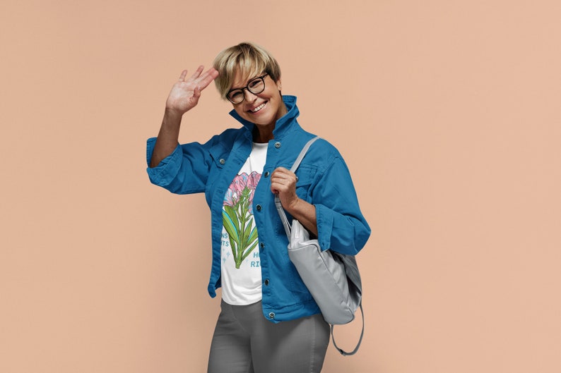 Puede incluir: Una mujer con una chaqueta vaquera azul sobre una camiseta blanca con estampado floral. Sonr&iacute;e y saluda con la mano izquierda. Lleva un bolso gris sobre el hombro.
